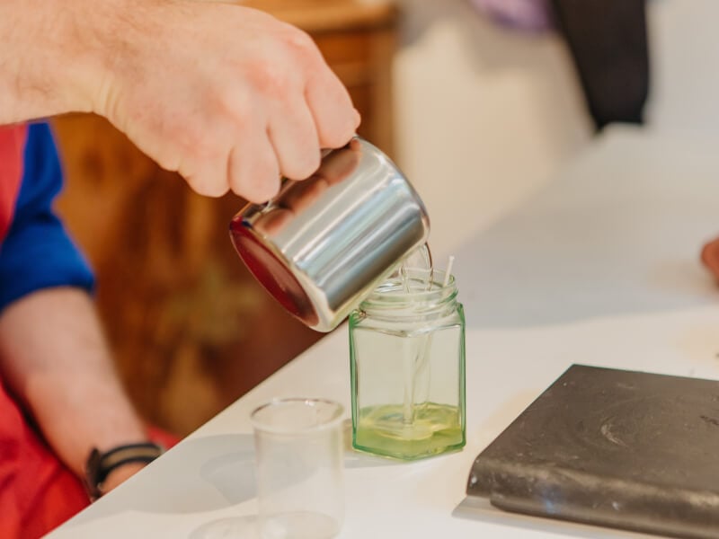 A man pours soy wax into a glass candle jar from a small metal jug